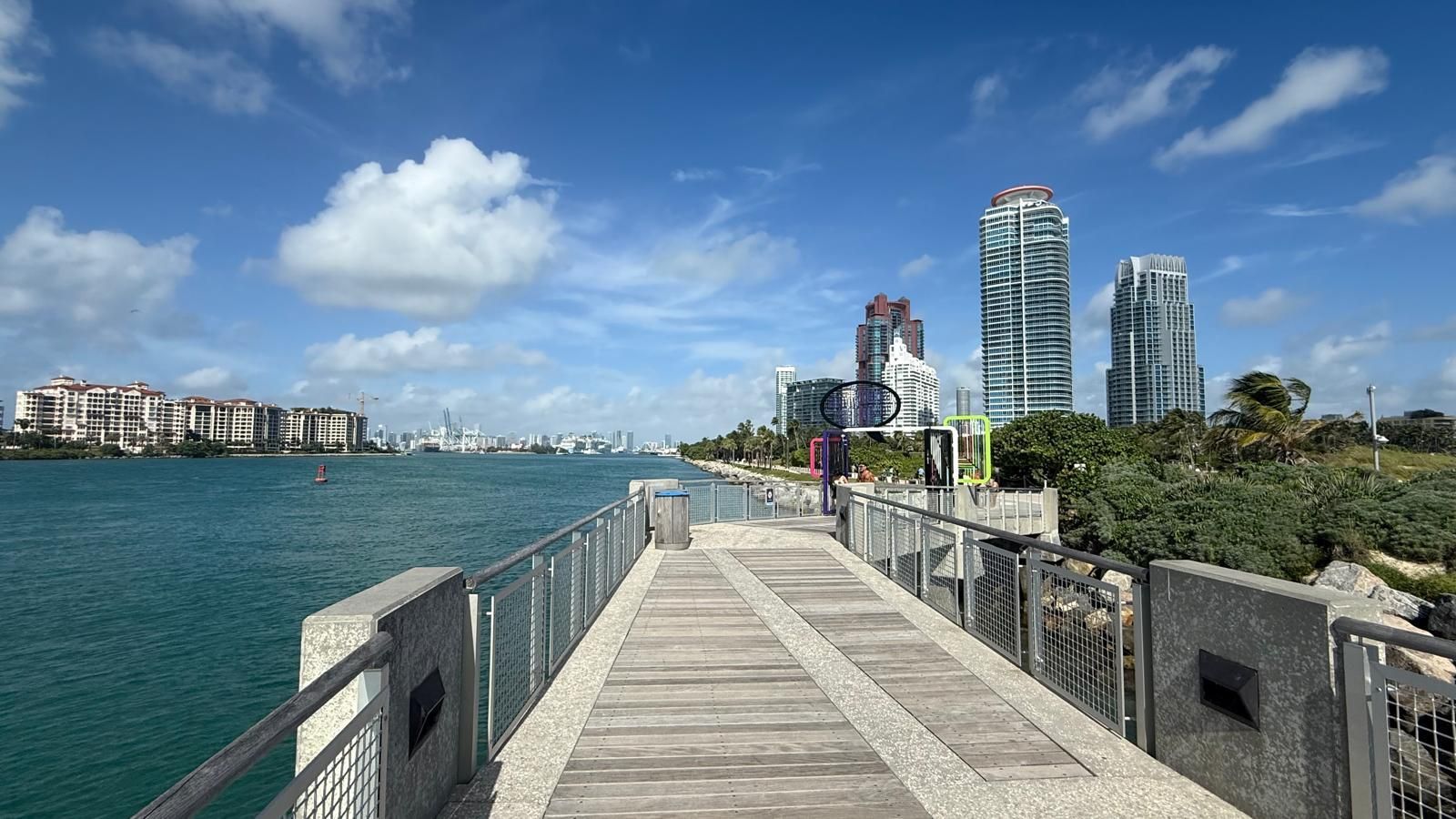 A wooden pier stretches toward the water with modern high-rise buildings in the background under a blue, cloudy sky.