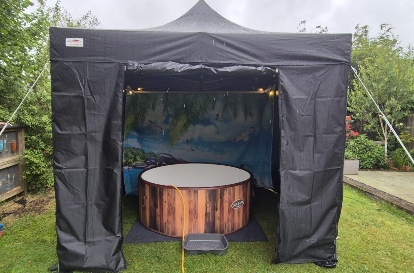 Black outdoor gazebo tent with a round wooden hot tub inside on grass.