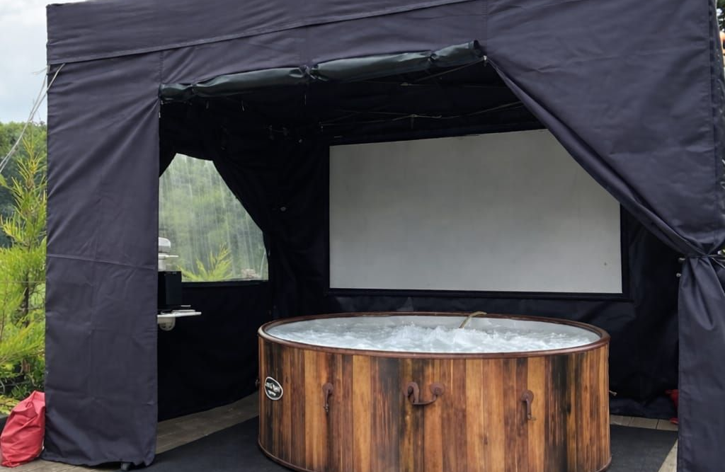 Black canopy tent with a round wooden hot tub and white screen inside on a paved surface.
