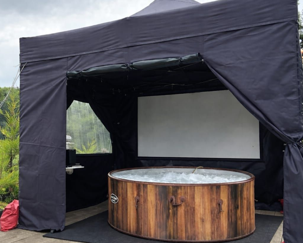 Black canopy tent with a round wooden hot tub and white screen inside outdoors