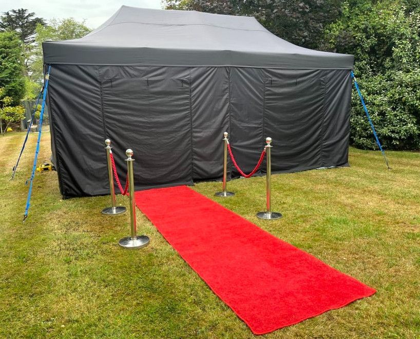 Red carpet leading to a black event tent on a grassy lawn, bordered by silver stanchions and ropes.