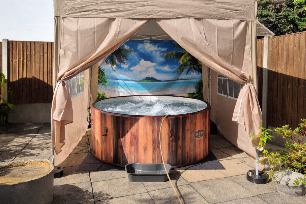 Round outdoor hot tub under beige gazebo with tropical mural backdrop and curtains in a patio yard