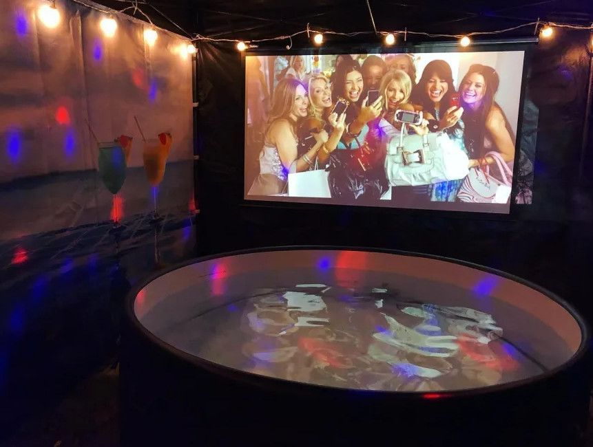 Dark room with a projected group photo and a round reflective floor lit by colorful string lights.