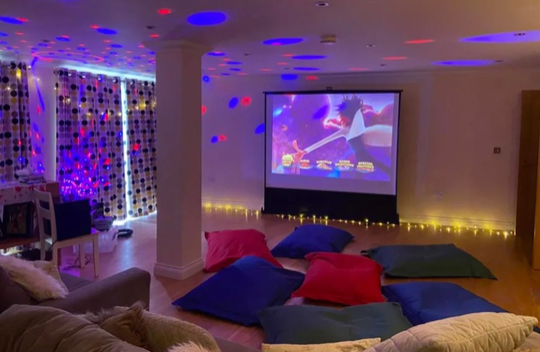 Cozy media room with colorful floor cushions, a projected movie, and string lights under a disco-style ceiling.
