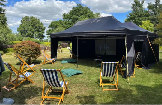 Black event tent on a grassy lawn with striped deck chairs and folding chairs under a blue sky.
