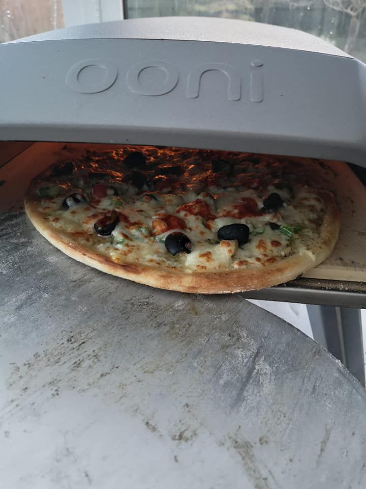Pizza baking in a gray Ooni oven, topped with olives and other ingredients.