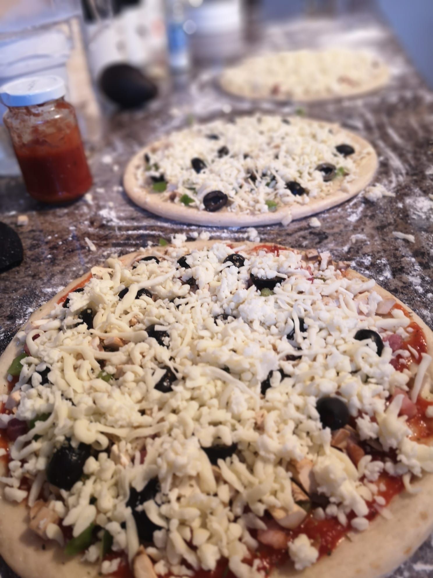 Two uncooked pizzas topped with cheese and black olives on a table, with a jar of tomato sauce nearby
