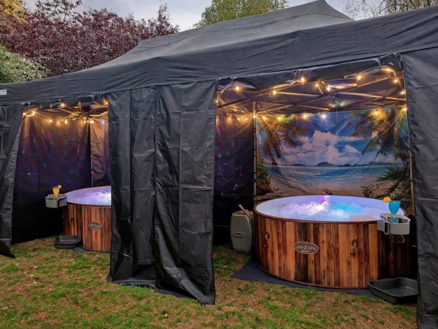 Two lit wooden hot tubs under a black canopy tent with string lights at dusk
