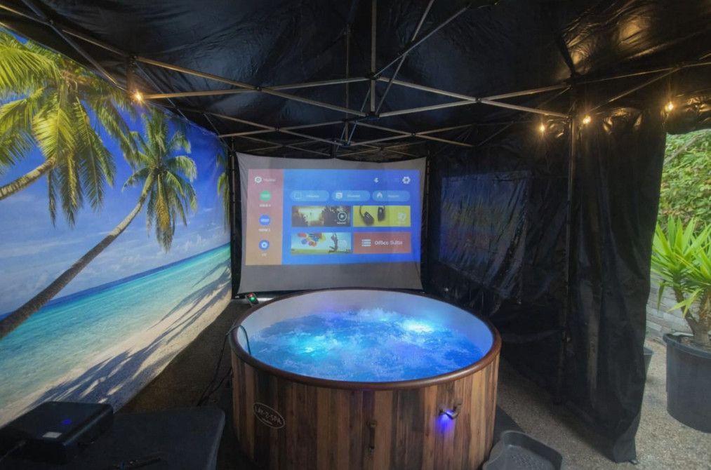 Tropical-themed booth with a glowing blue hot tub and a wall-mounted screen inside a black tent.