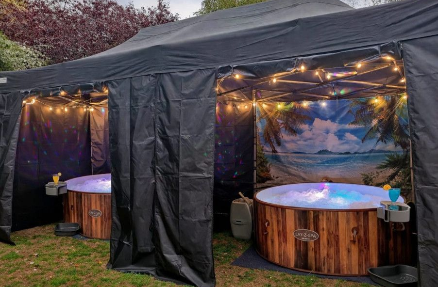 Black tent with string lights and two wood-sided hot tubs glowing blue and purple at dusk
