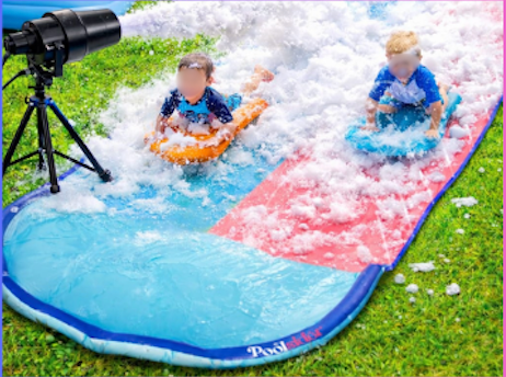 Two kids on orange and blue tubes sliding on a backyard water slide beside a sprinkler on grass
