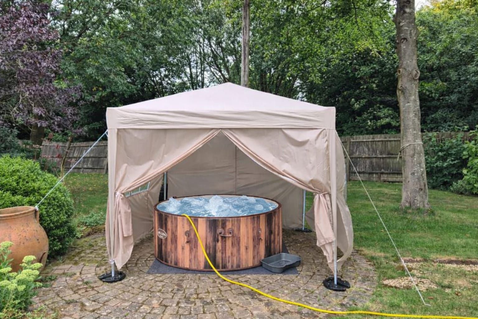 Beige outdoor gazebo with a round wooden hot tub in a backyard garden.