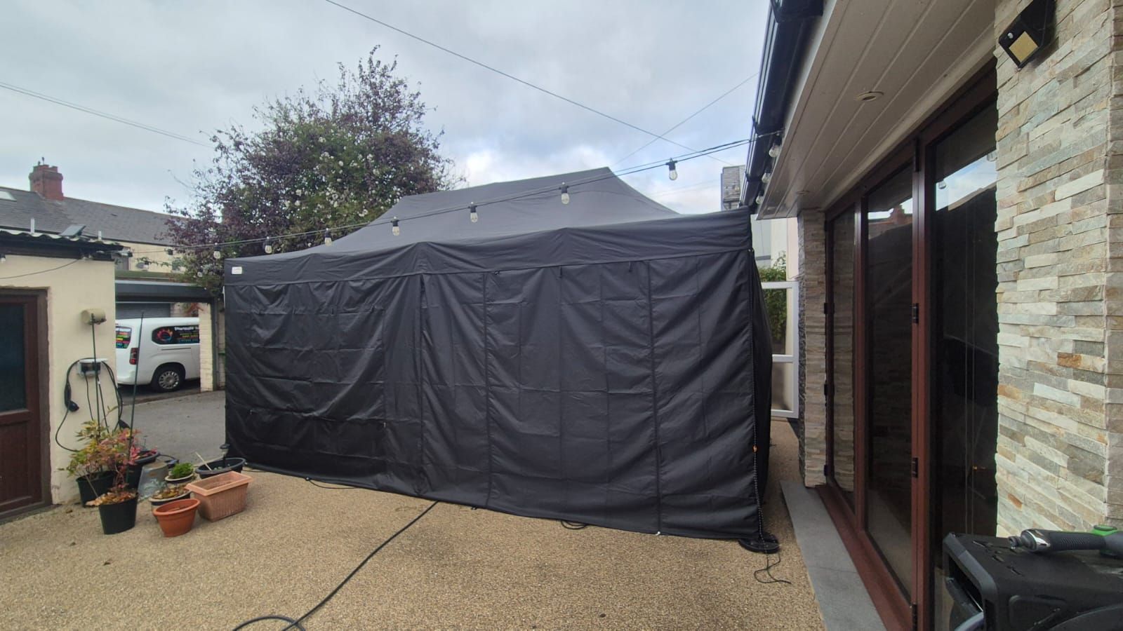 Gray outdoor canopy tent beside a stone house on a concrete patio, with potted plants nearby.