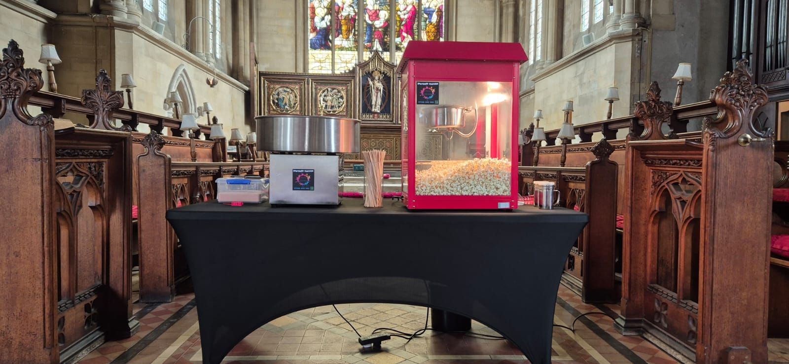 Red popcorn machine on a table inside a church-like hall with carved wooden pews and stained glass.