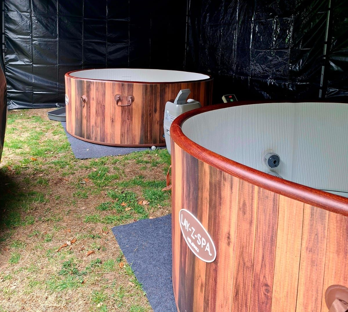 Two wooden hot tubs outdoors, one with water inside, on grass near a black tent backdrop