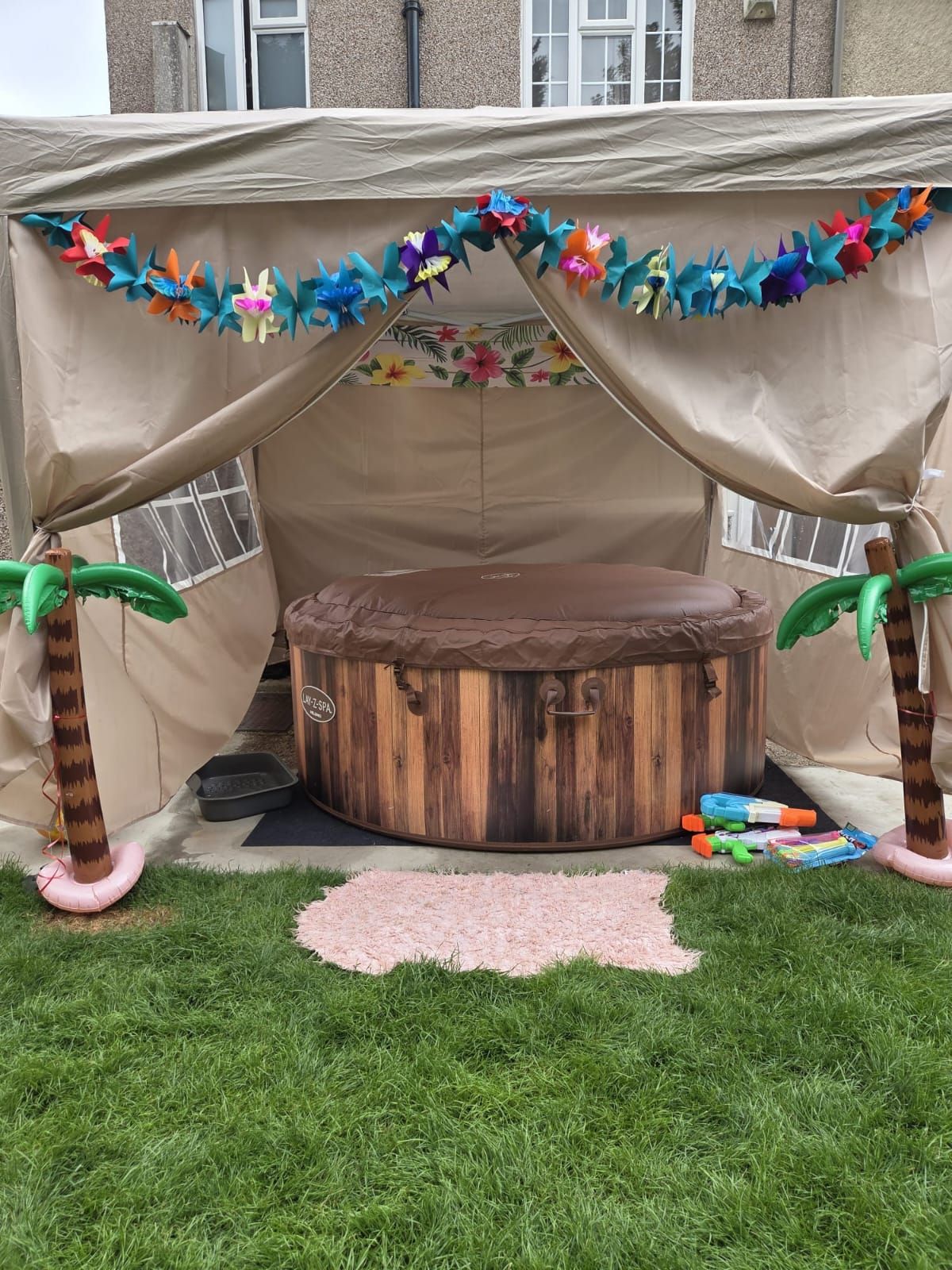 Decorated outdoor tent with colorful garlands, wooden barrel table, and palm-tree props on a lawn