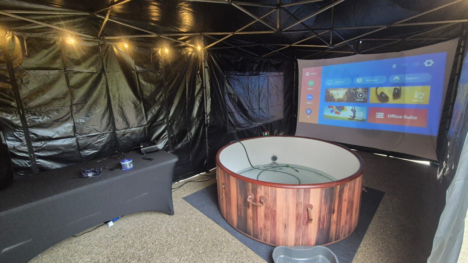 Backyard hot tub under a black tent, with a large outdoor screen displaying a game menu.