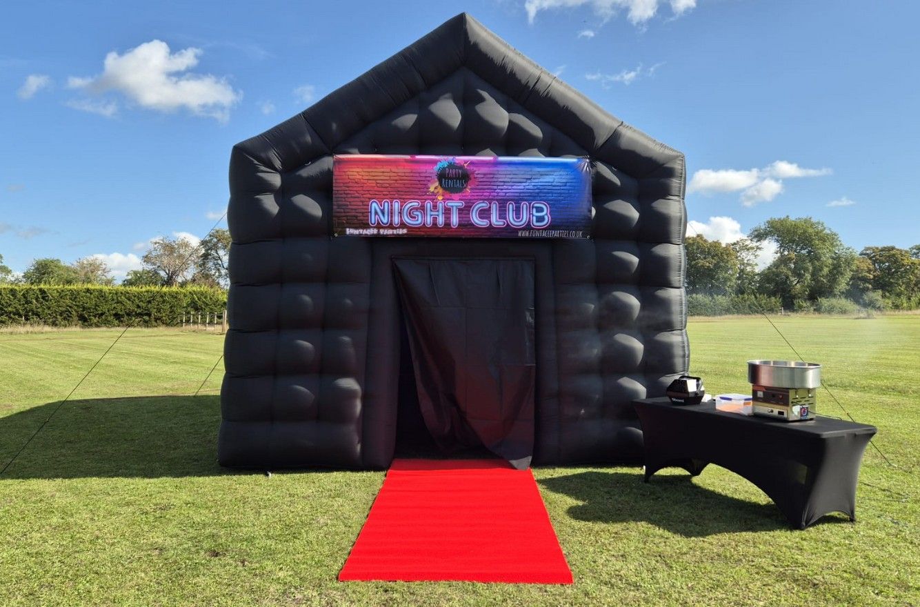 Black inflatable party tent with “Night Club” sign, red carpet, and table on a grassy field under blue sky