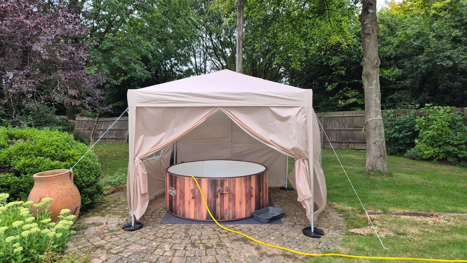 Small beige hot tub under a canopy tent in a grassy backyard with trees and a clay pot nearby