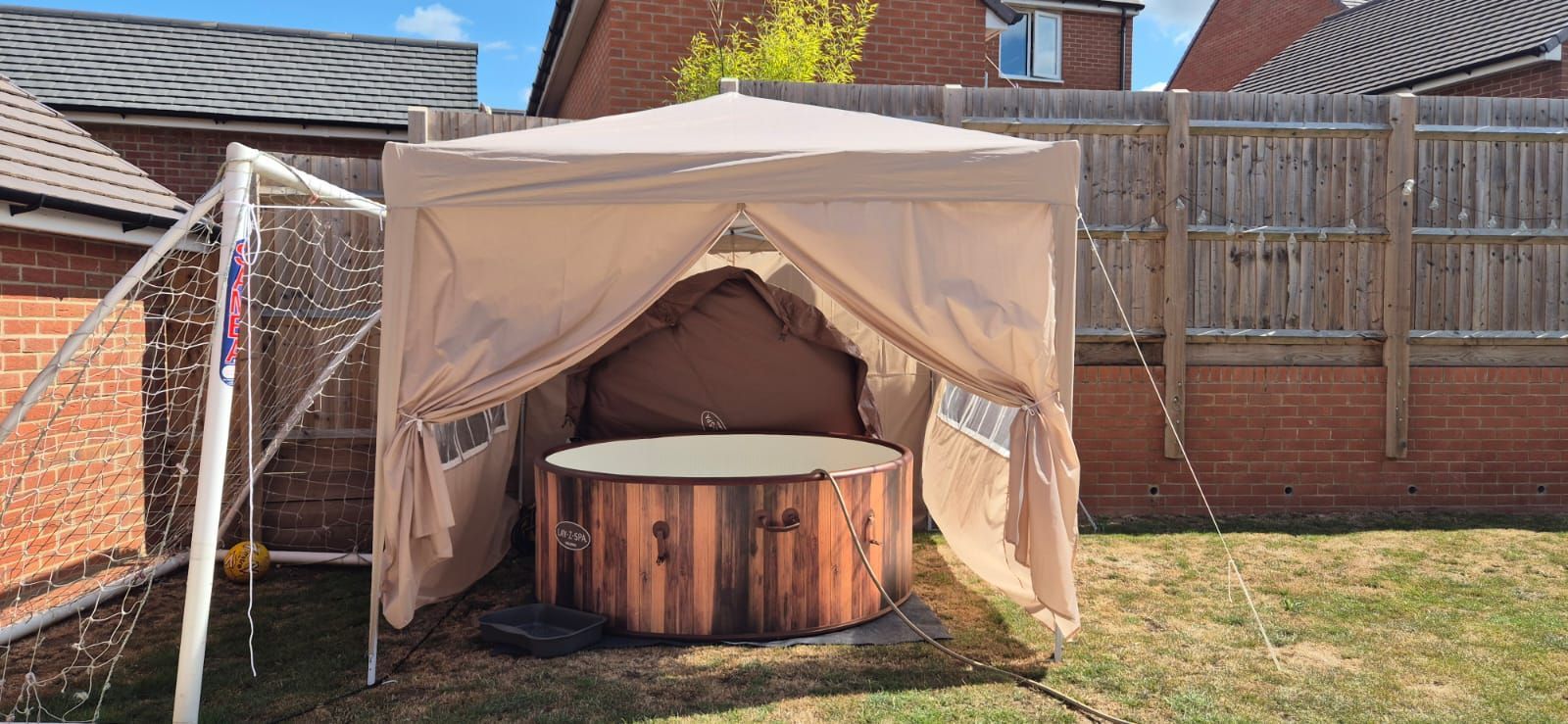 Beige canopy sheltering a round hot tub in a backyard beside a soccer goal and brick wall