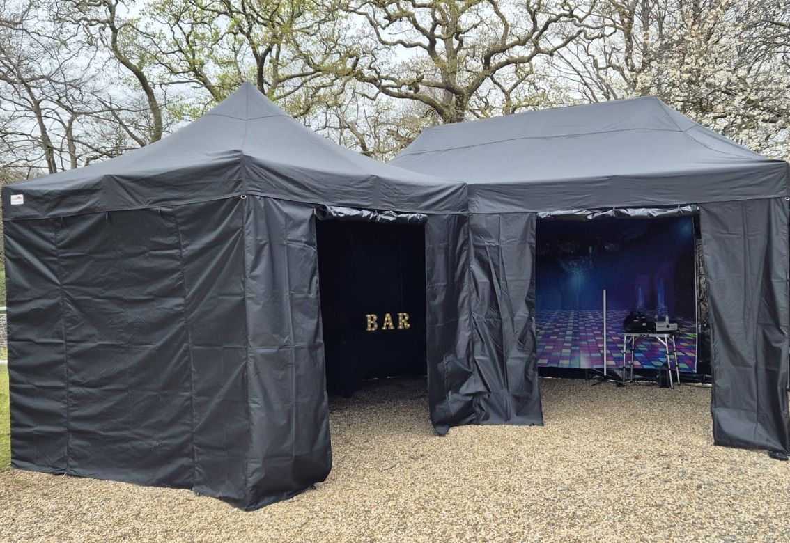 Black outdoor event tents with a bar sign and a small stage inside on a gravel lot under trees