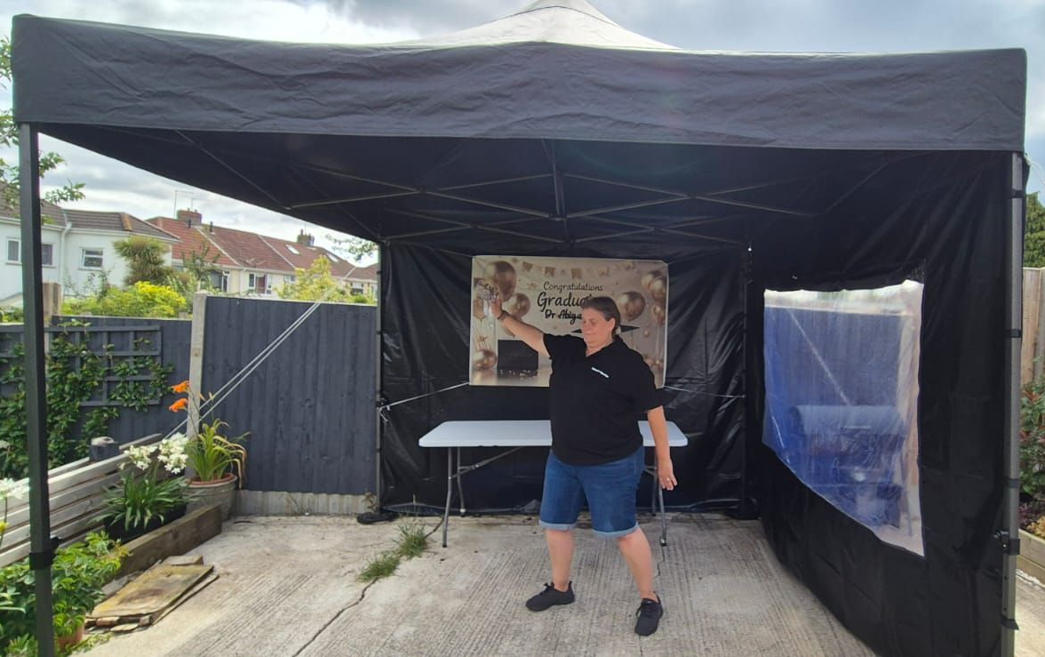Person standing under a black canopy tent in a driveway, gesturing toward a display board.