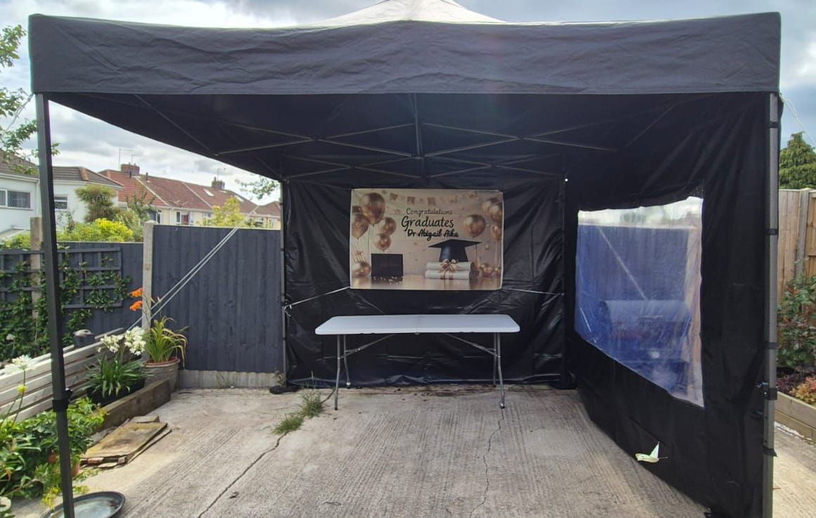 Black outdoor tent setup with a table and banner on a driveway, with clear side panels and garden plants nearby