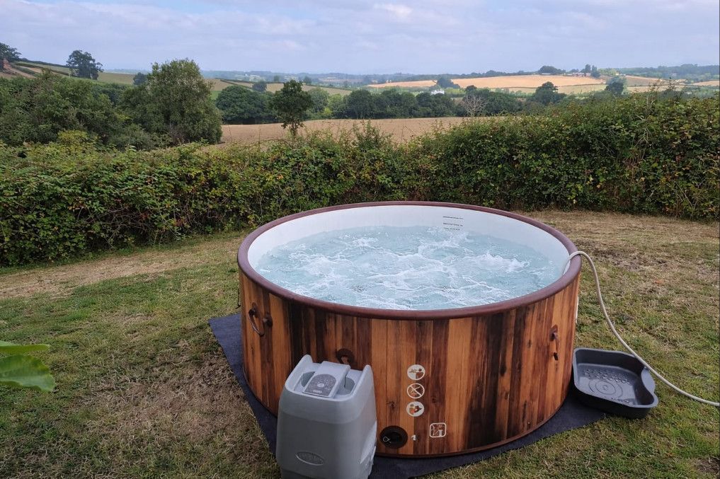 Round wood-fired hot tub in a grassy field, with hedges and countryside beyond.