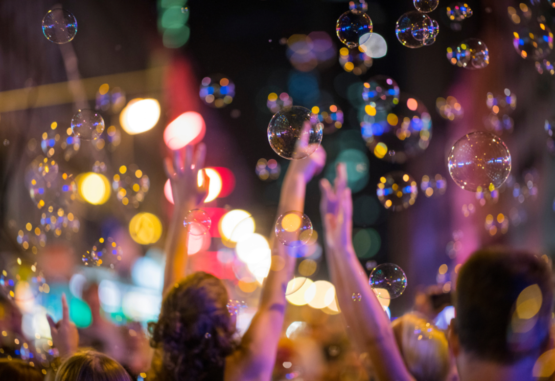 Crowd at a colorful concert or festival, with raised hands and floating bubbles in blurred lights