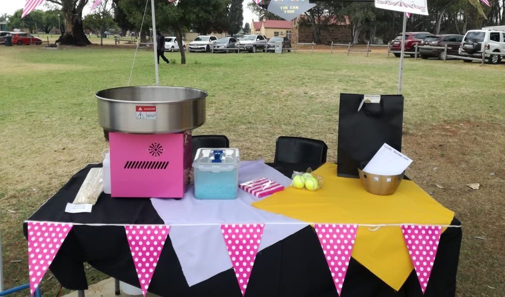 Outdoor dessert table with a pink cotton candy machine, bowls, and pink-yellow bunting on a grassy field