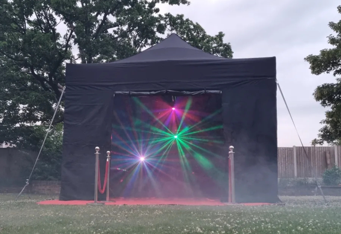 Black outdoor tent with colorful laser lights glowing at the entrance on a grassy lawn.