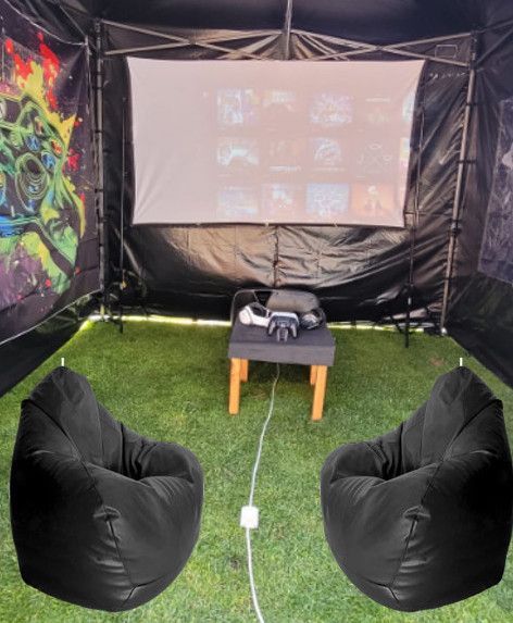 Two beanbag chairs facing a projector screen inside a black tent on grass, with a small table and projector.