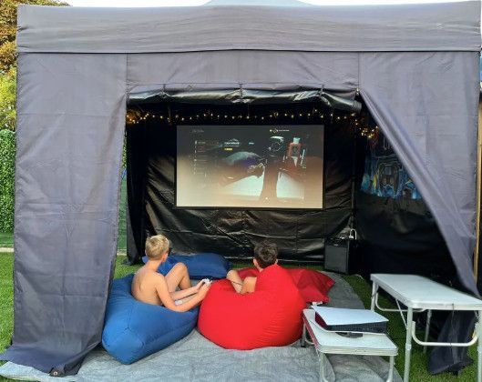 Two people sit on beanbags in a gray outdoor movie tent, watching a screen with tables beside them.