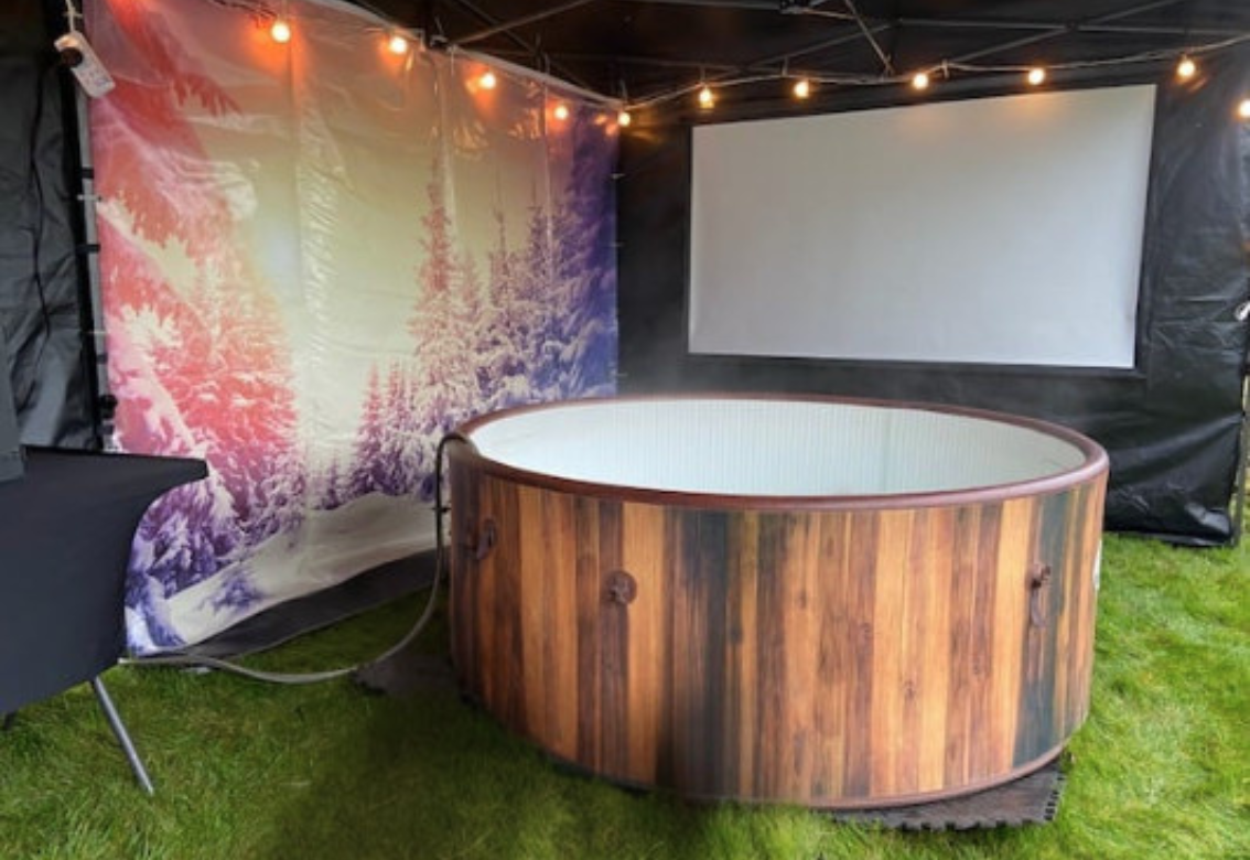 Outdoor hot tub lounge with wooden barrel hot tub, screen, and string lights under a tent