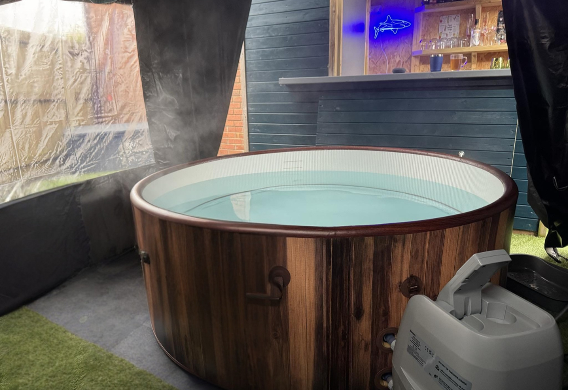 Wood-paneled hot tub filled with water on a patio with a grill and blue-lit bar in the background