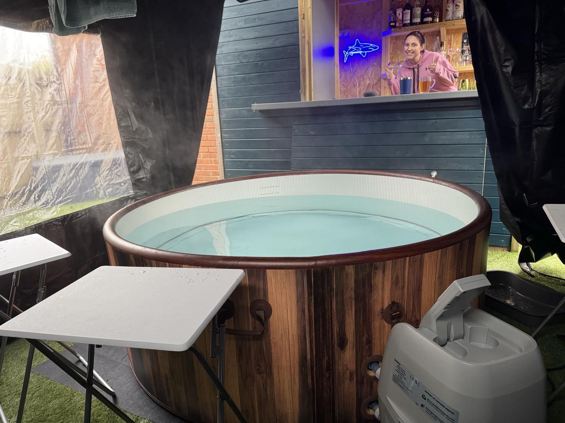 Round outdoor hot tub in a covered patio beside a small table and portable heater, with curtains and a window.
