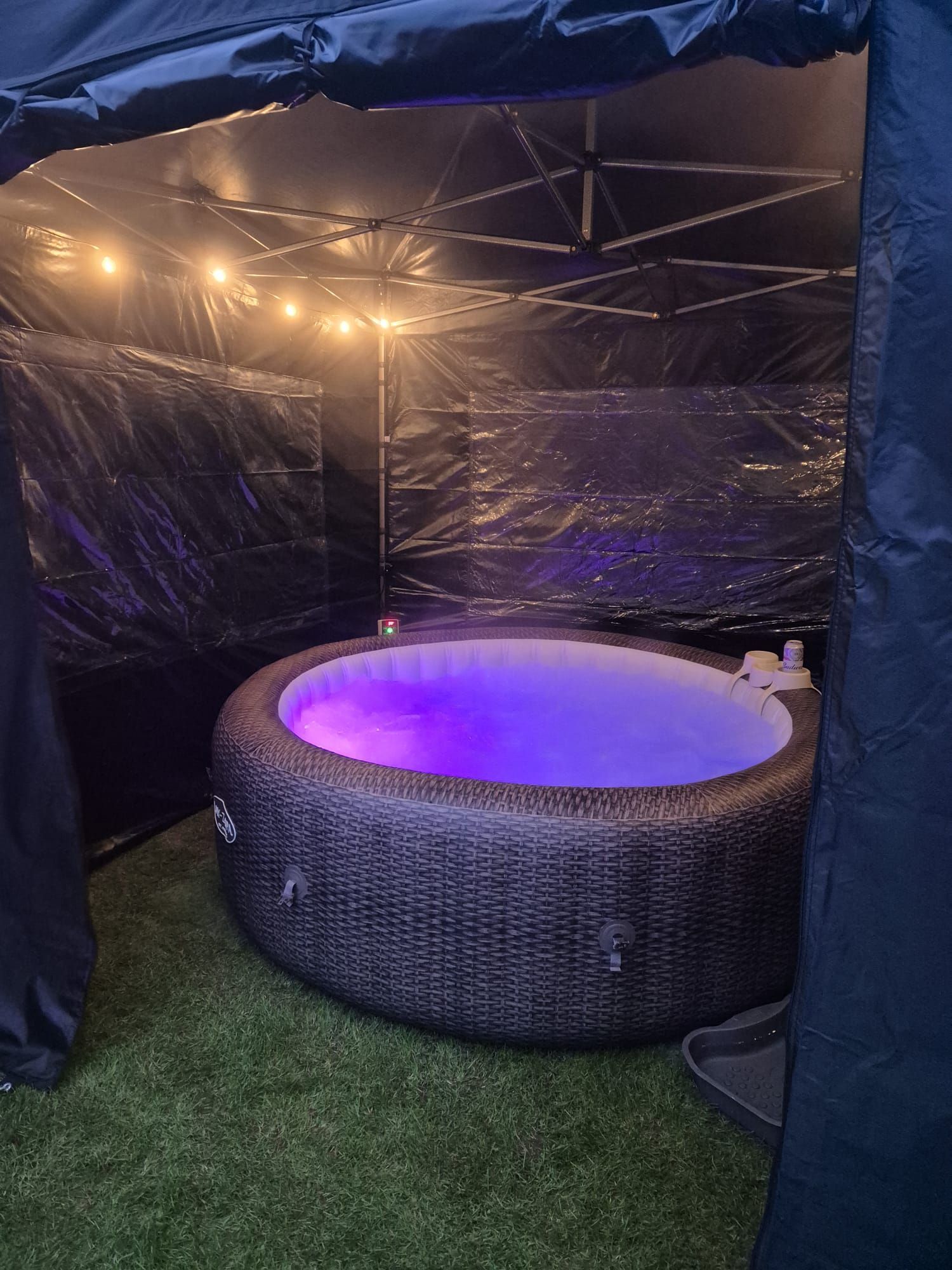 Round purple-lit hot tub inside a black canopy tent on artificial grass