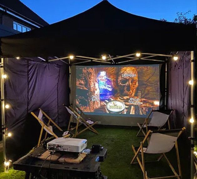 Outdoor movie setup under a black canopy with folding chairs, projector, and a projected scene at dusk