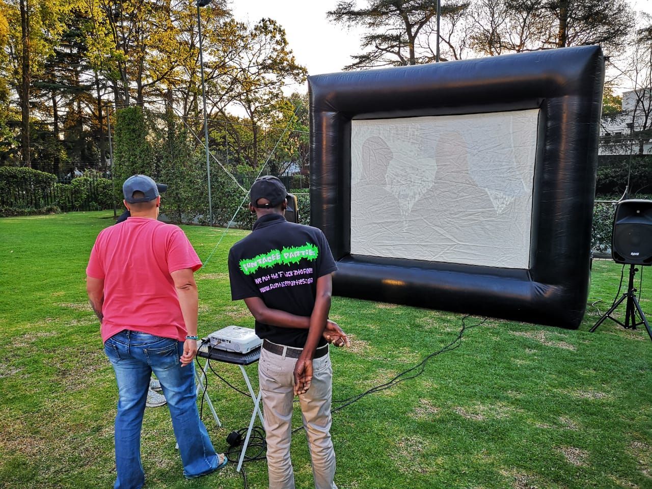 Two people stand on grass facing a large black inflatable screen outdoors.