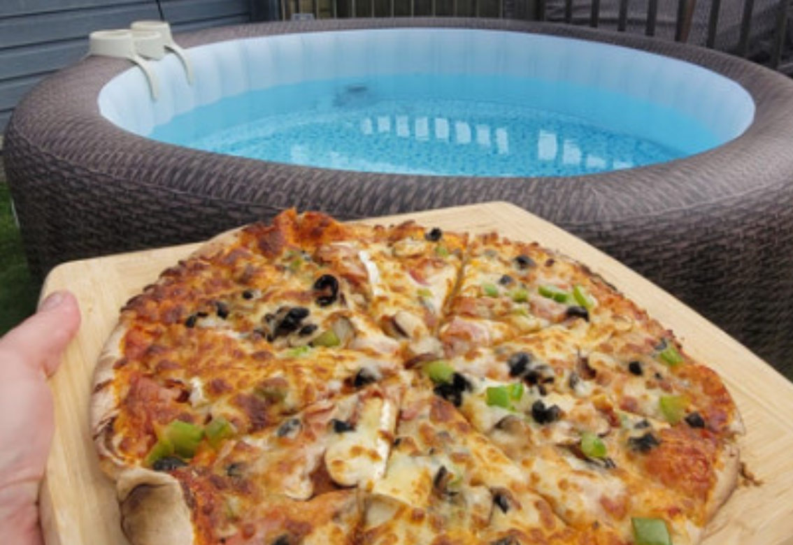 A hand holds a pizza in front of a hot tub with blue water and brown wicker sides.