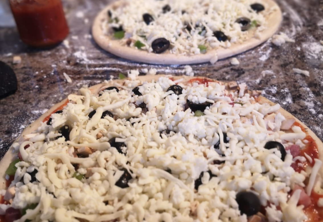 Two uncooked pizzas topped with shredded cheese and olives on a flour-dusted countertop