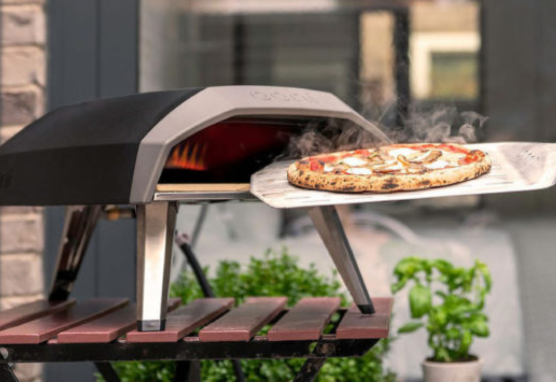 Pizza baking in a tabletop outdoor oven with one pizza on a peel, in a patio setting