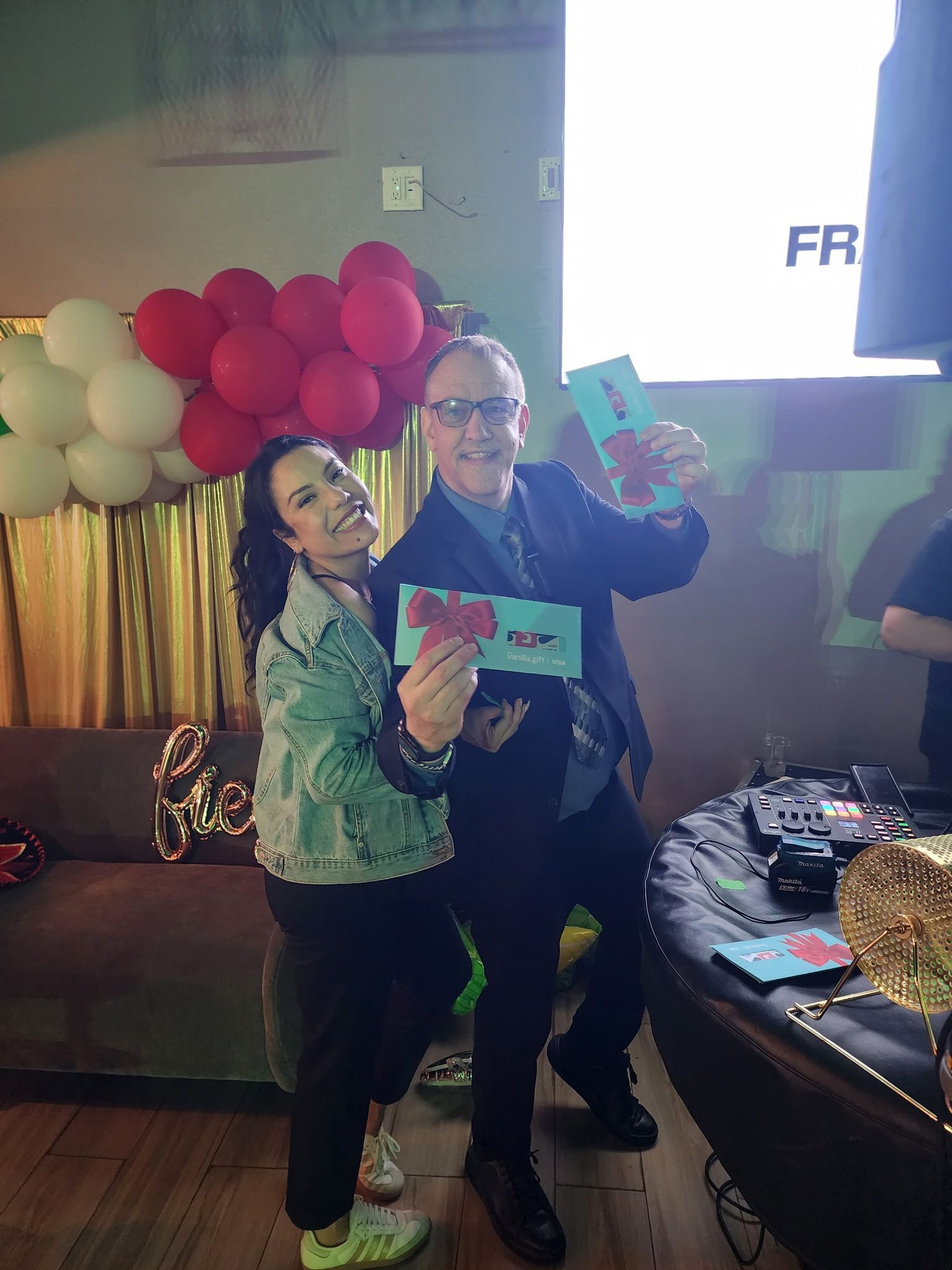 Two people pose indoors holding colorful cards, with red and white balloons and a TV in the background.