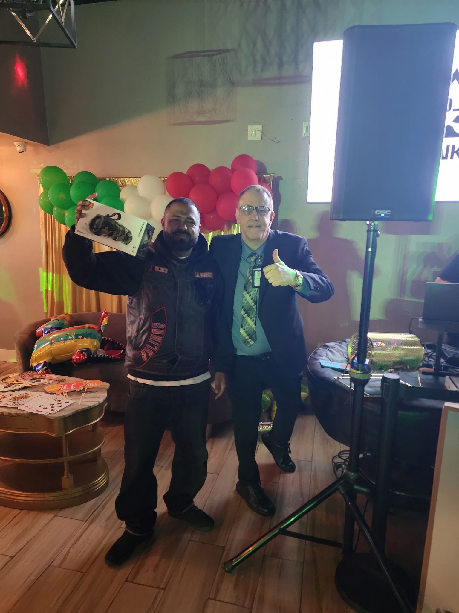Two people posing at a party with balloon decorations and a cake near a speaker stand