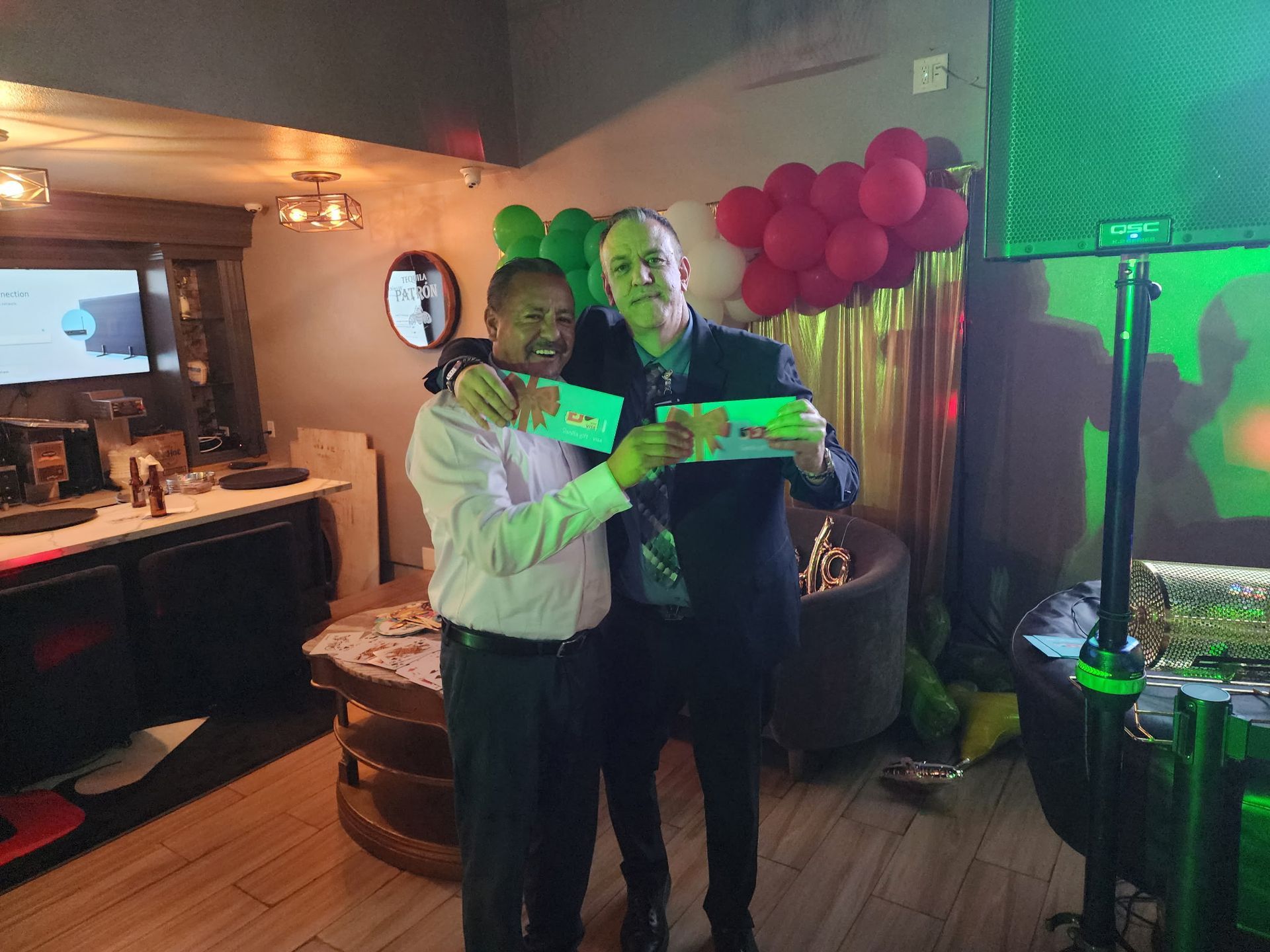 Two people hold a green certificate at a party with pink balloons and green lighting.