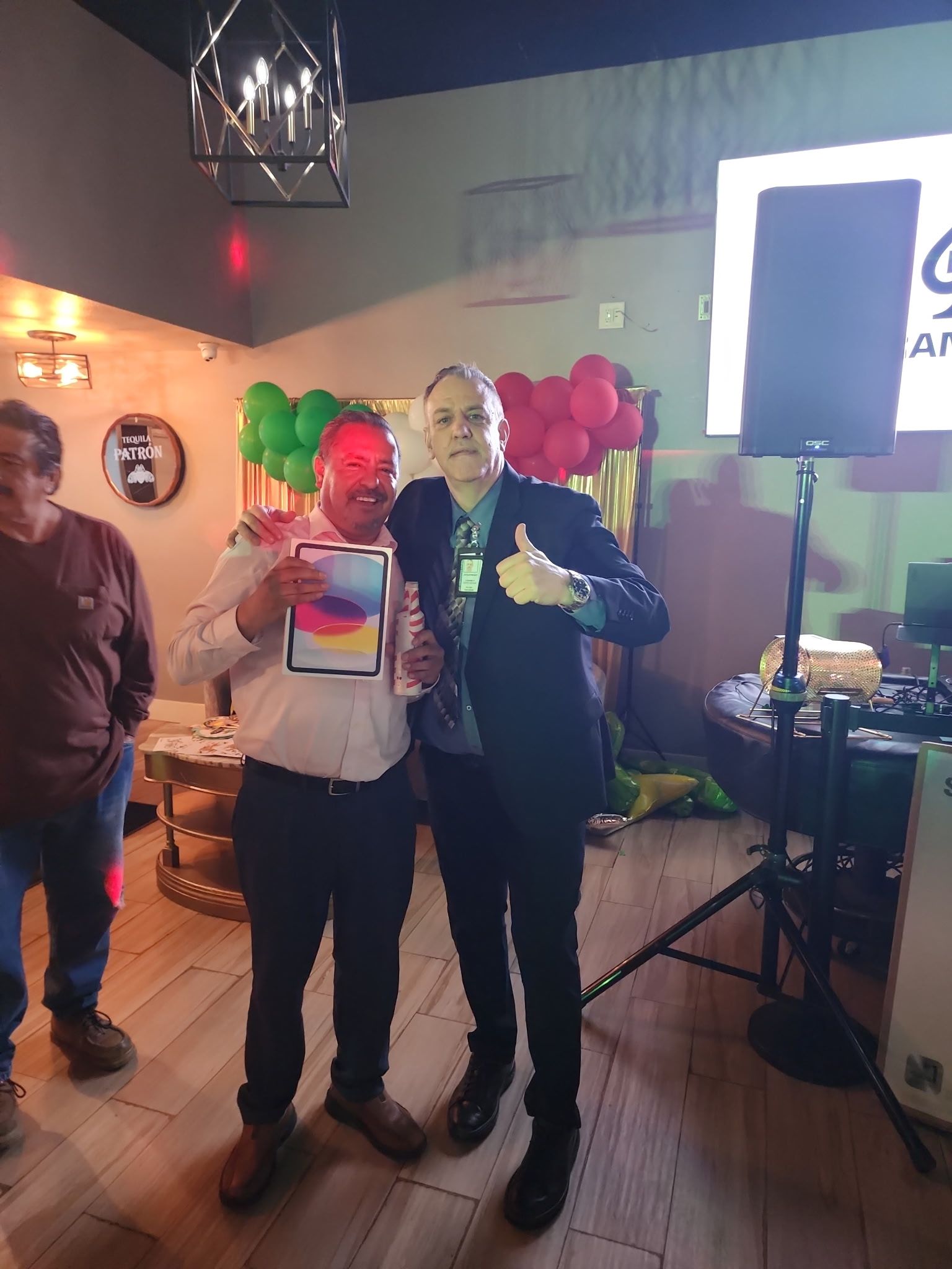 Two people hold a certificate and trophy at a party, with colorful balloons and a bright light in the background.