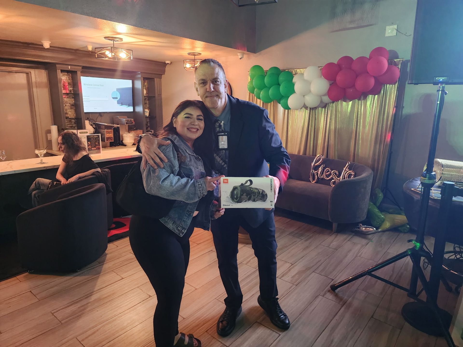 Two people posing in a decorated room with green and pink balloons, holding a framed sign and smiling