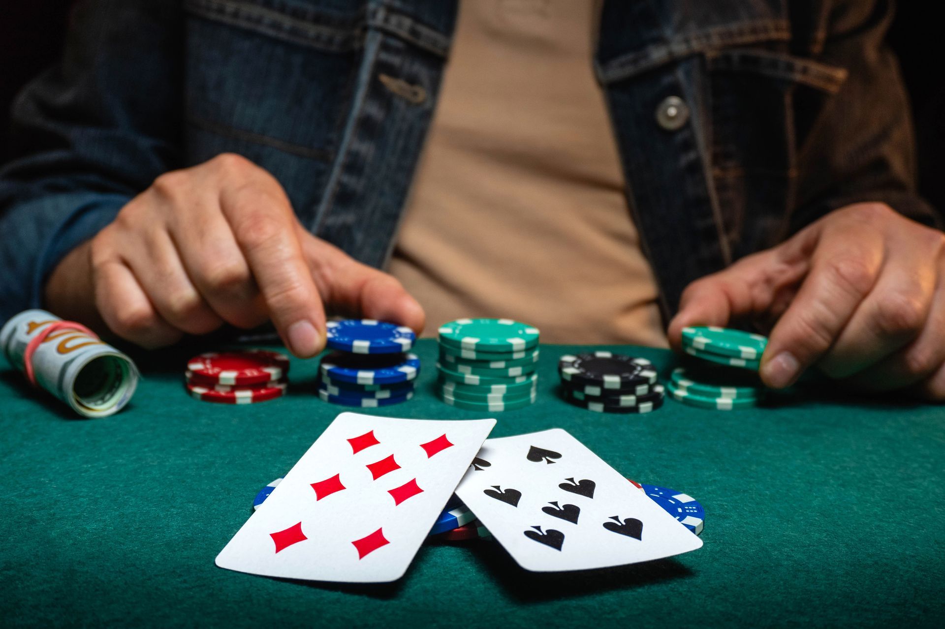 Close-up of a player's hands holding poker cards beside stacked chips and cash on a green felt table