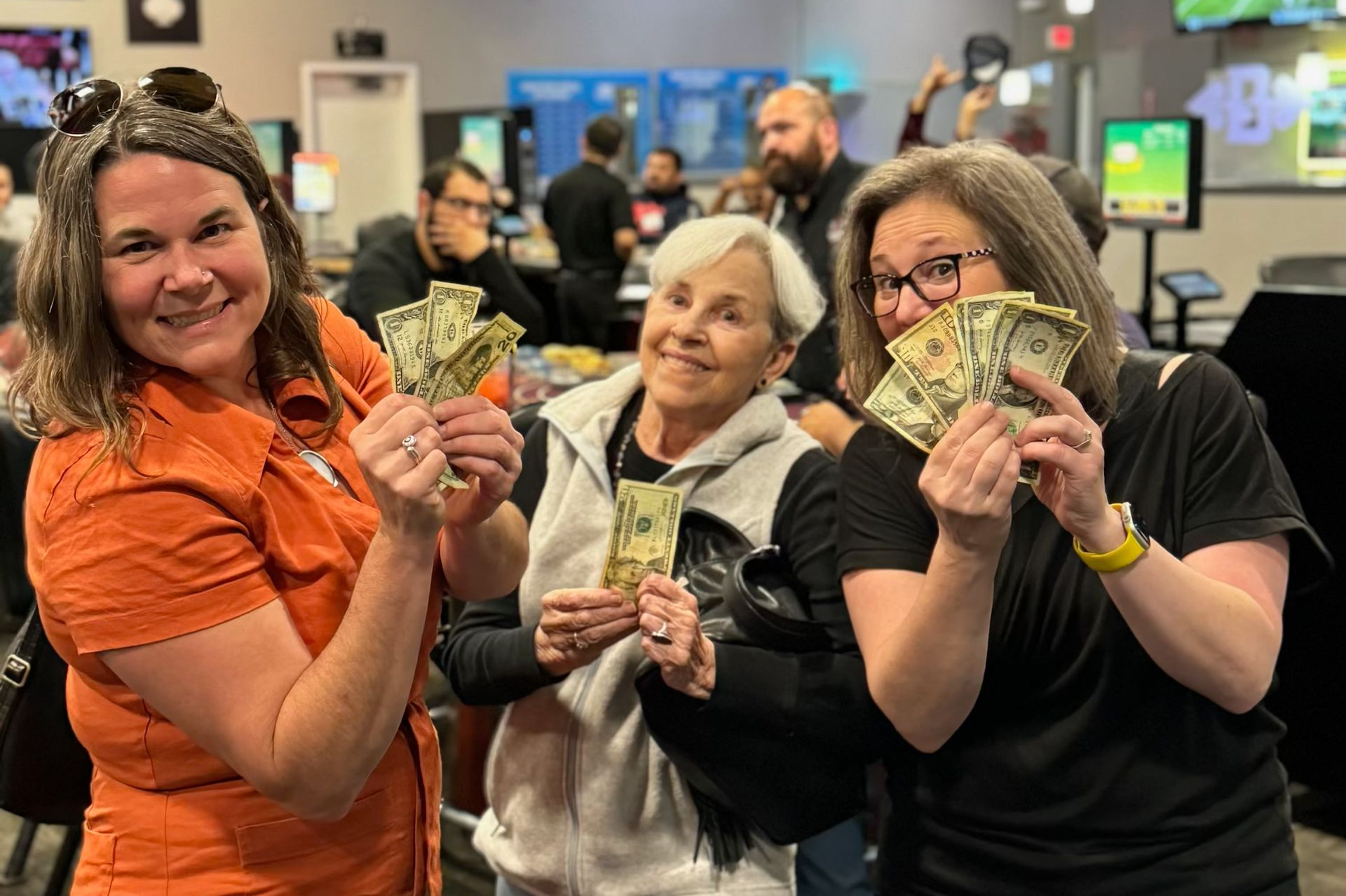 Three people in a busy indoor venue smiling and holding up cash prizes.