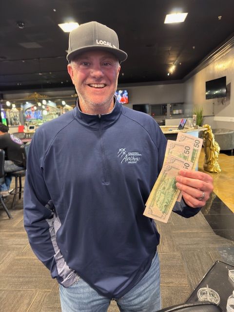 Man in navy jacket and cap smiling and holding a large cash prize indoors